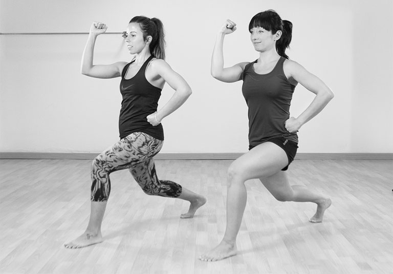 Dues dones fent zancada amb barra, entrenament Fit Dona
